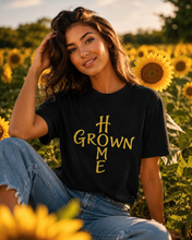 Carica l'immagine nel visualizzatore di Gallery, A woman sitting in a sunflower field wearing a black graphic t-shirt with 'GROWN ME' printed in yellow, paired with blue jeans.
