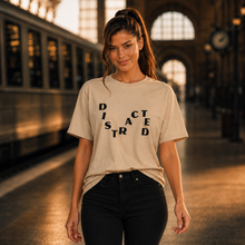 Ladda upp bild till gallerivisning, A woman stands on a train platform wearing a beige graphic t-shirt with the word 'DISRACTED' spelled out in black letters, paired with black pants.
