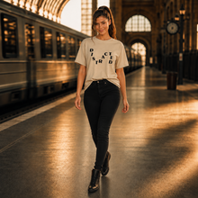 Ladda upp bild till gallerivisning, A woman walking on a train station platform wearing a beige graphic t-shirt with the word 'DISCRED' printed in black, paired with black pants and black boots.
