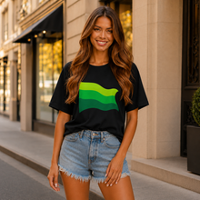 Load image into Gallery viewer, A woman stands on a city sidewalk wearing a black short-sleeve T-shirt with a green and yellow wavy stripe graphic on the front, paired with light blue denim shorts.
