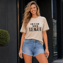 Load image into Gallery viewer, A woman wearing a beige graphic t-shirt with the text 'OUT OF YOUR LEAGUE' in black, paired with light-wash denim shorts, standing in front of a modern dark building.

